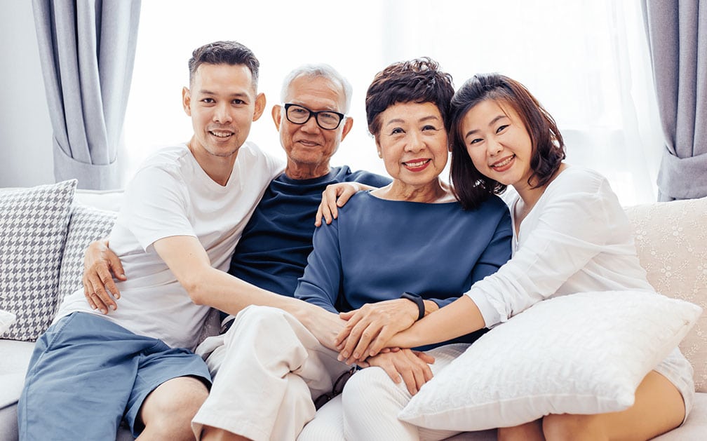 An Asian American family of four; a mother, father, son and daughter, sit on a couch together. The image is meant to signify how important assigning a life insurance beneficiary is to one’s family.