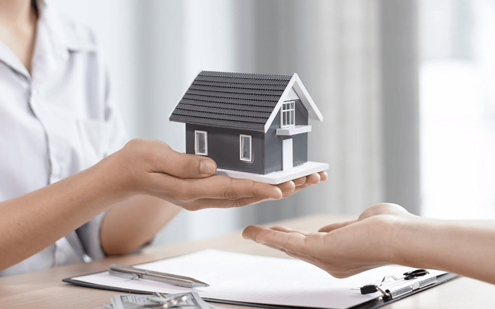 The image is of a small house held in two hands, with a receiving hand reaching for it. A clipboard with paper and pen resting on it is on the table below the hands. The image is used to signify the security mortgage protection insurance provides