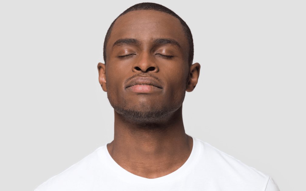 A photograph of a black man in a white t-shirt, showing him from the shoulders to the top of his head. His eyes are closed and his face is slightly raised upward in a relaxed pose. The image represents the benefits of mindfulness and relaxation
