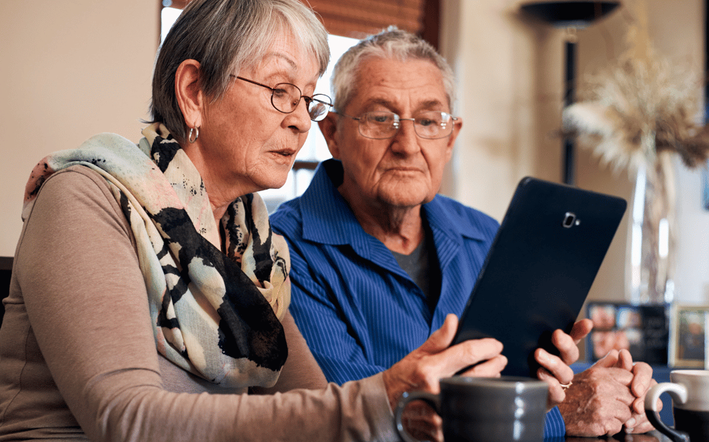The image features an older couple sitting at a table in their home looking at an electronic tablet. There’s a cup of coffee in the foreground. The image is used to depict a couple shopping for the best life insurance for seniors