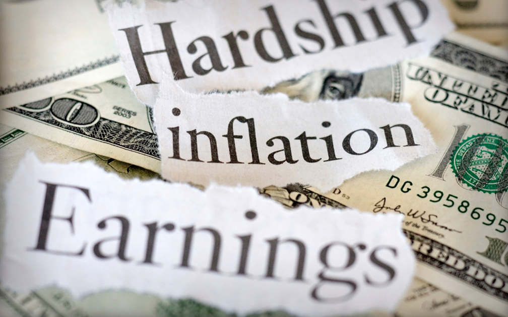 The image is filled dollar bills. Three ripped pieces of paper lie on top of them. On the piece of ripped paper at the top of the image is the typed word “hardship”. On the second ripped piece of paper in the middle of the image is the typed word “inflation”. And on the third ripped piece of paper at the bottom of the image is the typed word “earnings.”