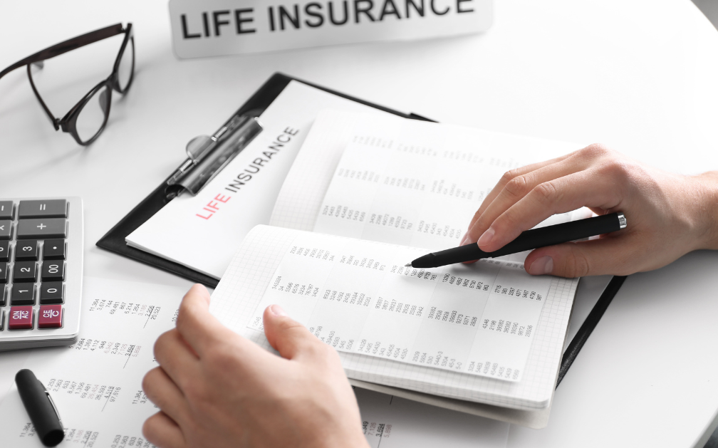 A desk conatains several items including a data sheet on which a black pen cover and calculator rest, a pair of glasses, and a folded card that reads “LIFE INSURANCE” in all capital letters. A clipboard with a paper that also says “LIFE INSURANCE” in all capital letters is under a booklet with more data. Two hands, one holding a black pen, hold the booklet.