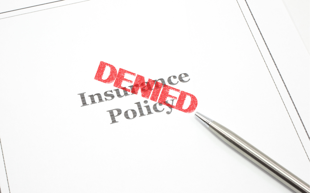 A piece of paper with the word “DENIED” in red ink stamped at a diagonal over the words “insurance policy.” A silver pen rests on a diagonal from the bottom right of the image.