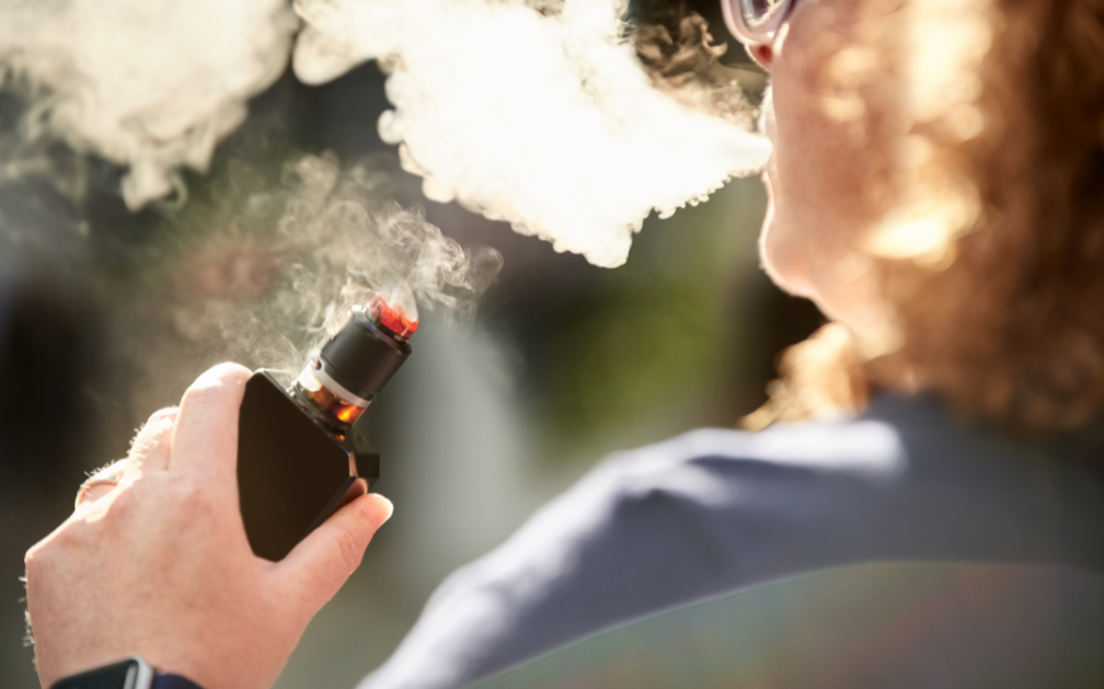 A woman with glasses and shoulder-length hair is blowing smoke from her mouth. She holds a black vaping device with a red tip in her hand. The image accompanies a blog post about if you can get life insurance if you smoke or vape.