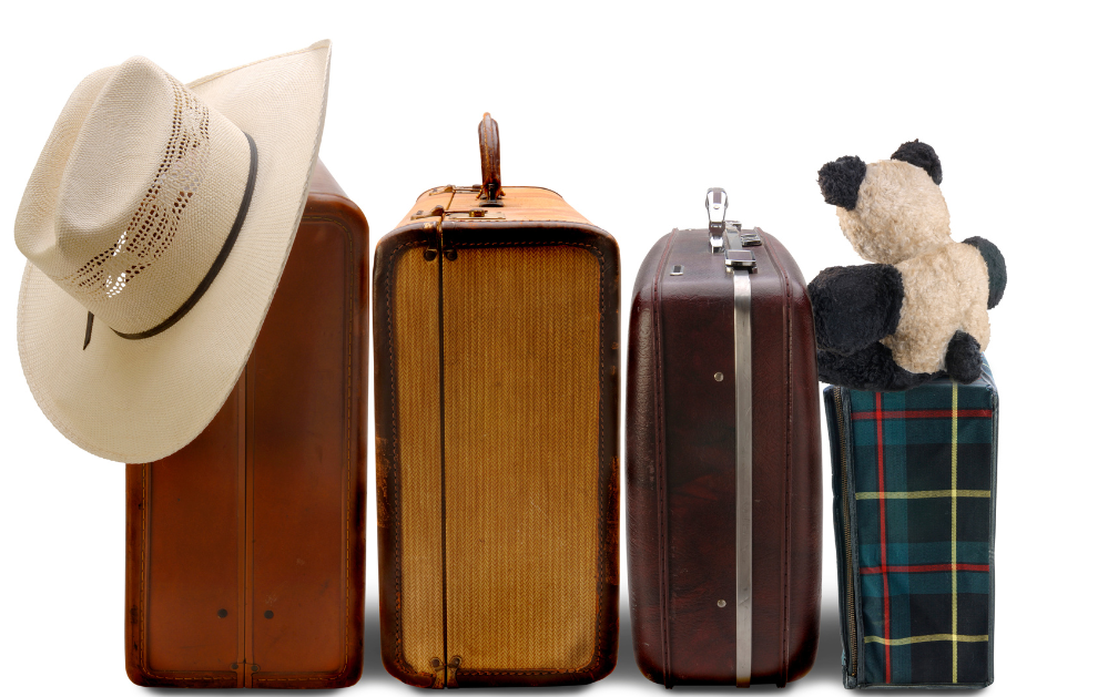 A row of four suitcases of different sizes, arranged from biggest to smallest, left to right. Resting on the biggest suitcase is a wide-brimmed hat. Resting on top of the smallest suitcase is a stuffed bear. The image accompanies an article about how moving overseas permanently impacts a U.S. life insurance policy.