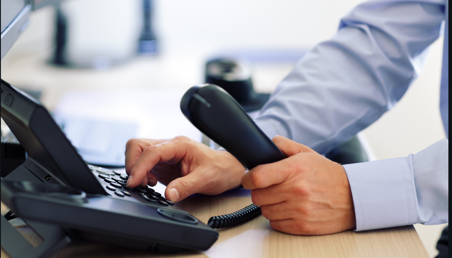 The image is of a man’s hands on a phone that is sitting on a desk. He is wearing long sleeves. One of his hands is dialing the phone and the other is holding the receiver. The image accompanies an article about why insurance companies conduct phone interviews.